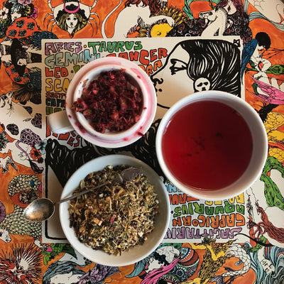 A tea cupping set with LUCID Tea Hibiscus Herbal Tea Blend on an orange tray decorated with colorful astrological art.